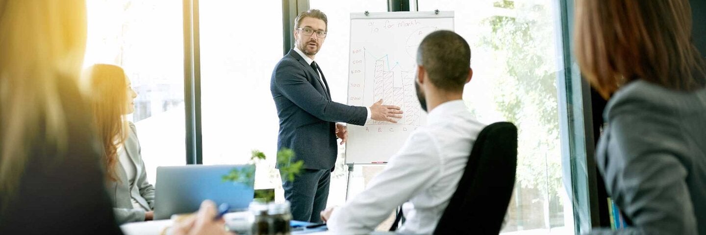 Man presenting business plan on whiteboard to room of coworkers.