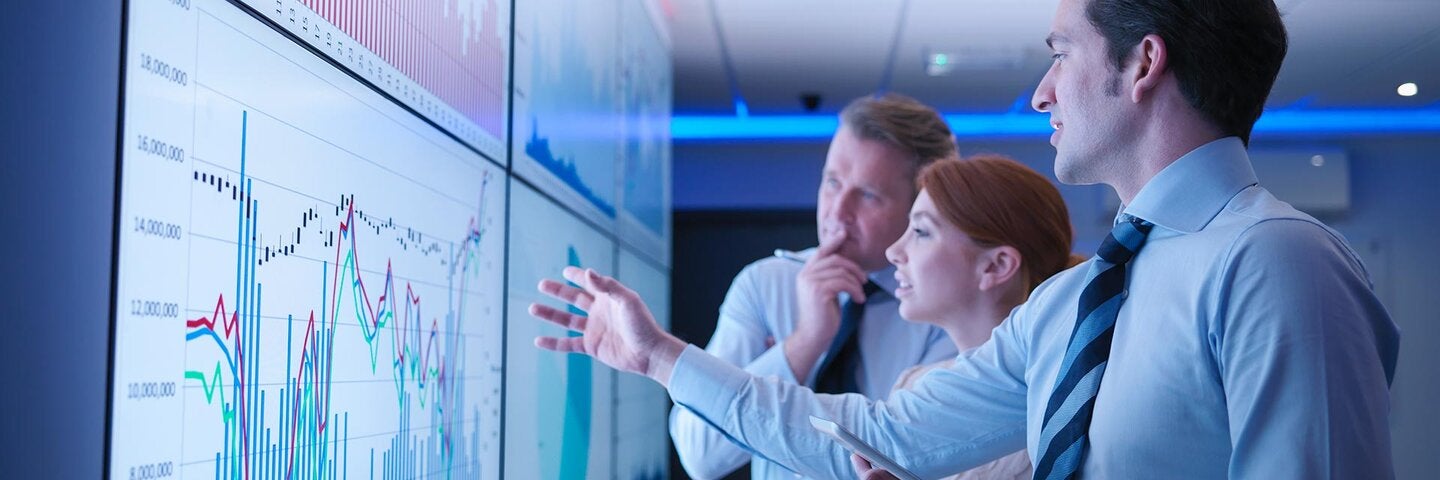 Three business people discuss graphs on screen in meeting room