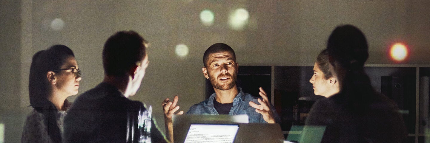 Four professionals collaborating around a lit table with laptops in a dimly lit office at night, with a man gesturing as he speaks to the group.